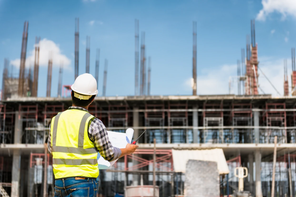 Chantier de construction en extérieur avec ouvrier en gilet haute visibilité jaune et casque blanc consultant des plans architecturaux