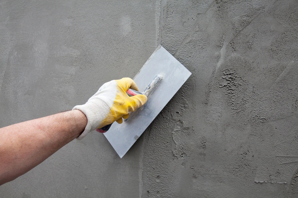 Ouvrier appliquant un enduit de finition sur un mur extérieur avec une taloche bleue, portant des gants de protection orange