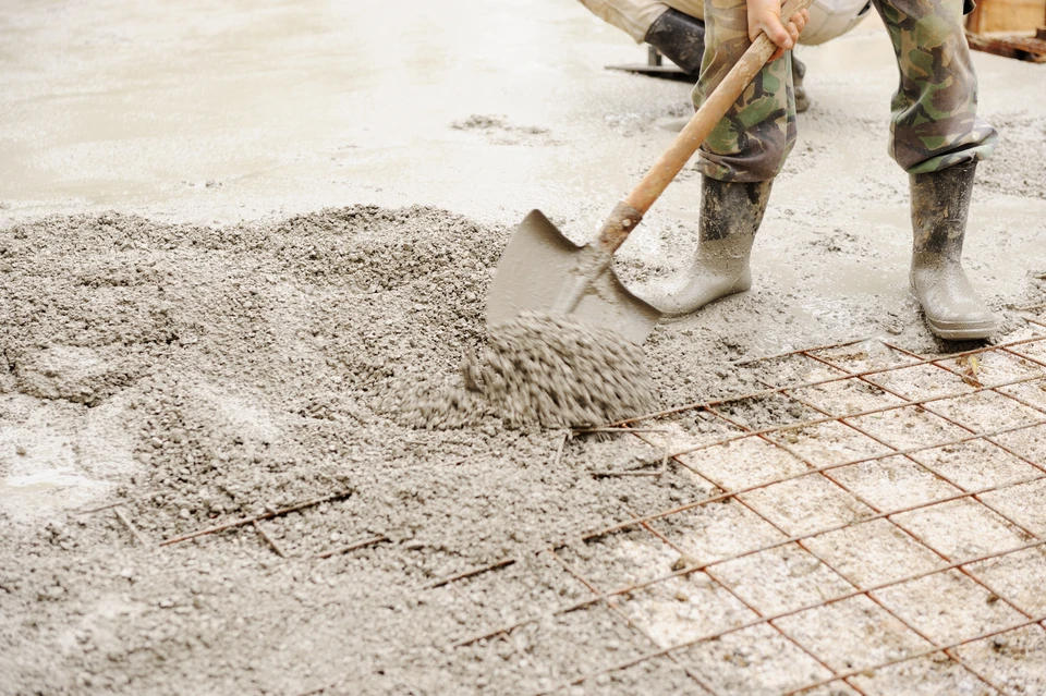 Photographie de chantier montrant un maçon expérimenté travaillant sur des fondations. La scène doit montrer clairement du travail de maçonnerie : un ouvrier en tenue de chantier complète (casque, gants) posant des briques ou des blocs de béton pour les fondations d'un bâtiment, avec des matériaux de maçonnerie visibles (briques, mortier, niveau). Le contexte doit être celui d'un vrai chantier de construction, mettant en avant l'expertise et la rigueur du travail de fondations.